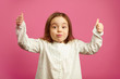 © Ilshat - Surprised little girl with raised hands, shows thumbs up, has a stunned face, expresses surprise and joy on pink wall