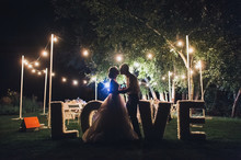 Beautiful Newlyweds Kiss Tenderly At A Wedding Party With Lamps. Stylish Wedding Ceremony. Love In The Frame. Designer Letters.