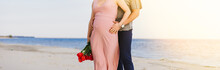 Pregnant Woman With A Bouquet Of Tulips Is On The Beach, And Her Husband Is Hugging From Behind.