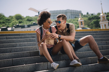 young male traveler trying to eat girlfriend croissant on stairs