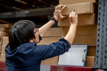 Wall Mural - Young Asian man doing stocktaking of product in cardboard box on shelves in warehouse by using clipboard and pen. physical inventory count concept