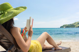 Fototapeta  - Side view of a young beautiful woman reading a book while sitting on a wooden lounge chair at the beach in a sunny day during summer vacation in Flores Island, Indonesia