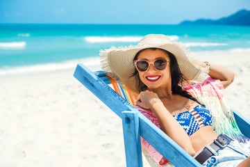 Wall Mural - Beautiful girl sitting on the beach by the sea in the color deck chair, dressed in a pair of straw hat with sunglasses, summer vacation trip, emotion joy happiness