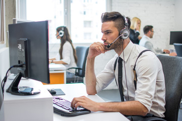 Wall Mural - Beautiful young man working in call center. Customer support operator with headset working in office with his colleagues.