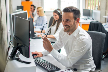 Smiling handsome office worker using computer in office with colleagues in the background.