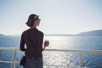 Wall Mural - Traveller girl standing on ferry boat, looking at the sea and holding a coffee cup, travel and active lifestyle concept