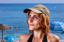 Young Woman At Beach, Summertime. Close-up Of Pretty Girl With Nose Ring And Wet Hair Under The Sun, In Front Of Parasols And The Blue Sea. She Wears A Green Baseball Cap And A Black Swimsuit