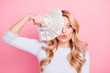 Portrait of playful foolish girl with curls, modern hairstyle, pout lips hiding half of her face with fan of much money isolated on pink background