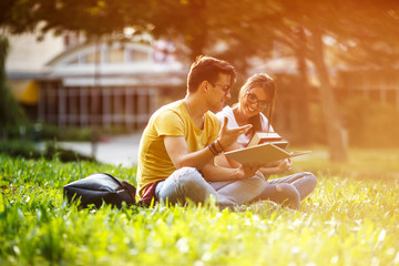 Two young students sitting at the campus yard , reading book and preparing for university exam.