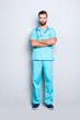 © deagreez - Full size body portrait of brutal virile man with stethoscope on his neck in blue lab uniform, holding his arms crossed, isolated on grey background