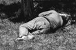 © Srdjan - Old barefoot homeless or refugee man sleeping on the grass in the park using his travel bag as pillow, social documentary street concept, dark black and white