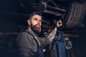 Bearded auto mechanic in a uniform repair the car's suspension with a wrench while standing under lifting car in repair garage.