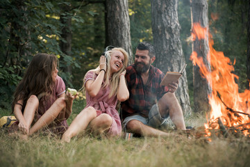 Wall Mural - Friends relax at bonfire flame with sparks in vintage style. People camping at fire in forest. Women and bearded man at campfire. Eating food, reading book and entertainment. Summer vacation concept