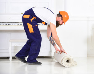 Wall Mural - Relocating concept. Man with beard, worker in overalls and helmet rolling carpet, white background. Loader wrappes carpet into roll. Courier delivers furniture in case of move out, relocation.