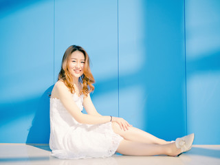 Wall Mural - Beautiful Chinese brunette woman in white wedding dress posing and sitting on floor with blue wall background. Sunny portrait of glamour young stylish lady. Emotions, beauty and lifestyle concept.