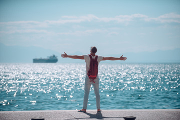 Wall Mural - man traveler with backpack hold rised hands at sea water. travel and wanderlust concept. summer vacation and adventures. young man is going to marine trip. futute life and freedom concept