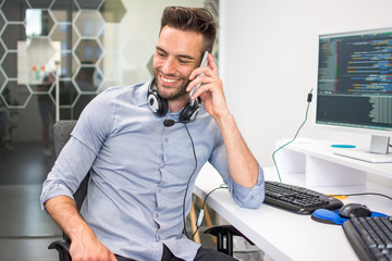 Wall Mural - Handsome programmer with headset talking on mobile phone at his workplace.