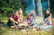 © Volodymyr - Group of friends camping in woods. Two couples cooking sausages over fire. Brunette and blond girls relaxing with their boyfriends outdoors