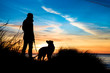 Silhouette von einem Mann und einen Hund am Strand bei Sonnenuntergang umrahmt von Gräsern