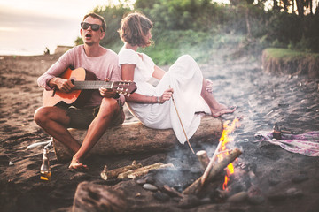 Guy with guitar and his girlfriend sitting on log and singing for his friends on summer evening by campfire