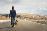 Fototapeta  - A stylish bearded hipster in sunglasses with a vintage backpack walks along the asphalt road on a sunny day. The concept of hitchhiking and hiking