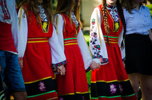 Unrecognizable Girls In Traditional Bulgarian Costumes With Red Dresses And Patterns On White Shirts Holding Hands