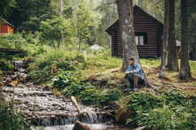 Stylish Traveler Man Sitting Near Cottage And River In Sunny Forest And Mountains. Travel And Wanderlust Concept. Space For Text. Happy Hipster Guy Relaxing In Woods. Summer Vacation