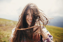 Happy Traveler Hipster Girl With Windy Hair And Smiling, Standing On Top Of Sunny Mountains. Space For Text. Stylish Woman Waving Hair. Atmospheric Moment. Travel And Wanderlust