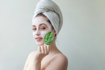 Wall Mural - Beauty portrait of a smiling brunette woman in a towel on the head with white nourishing mask or creme on face and green leaf in hand on white background isolated. Skincare cleansing eco organic