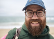 © Cavan Images - Close-up portrait of smiling bearded man wearing cap and eyeglasses at beach