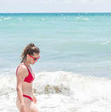 Fototapeta  - Young happy smiling woman standing in red swimsuit bikini bathing suit, sunglasses looking down at wave on Hollywood, Miami Beach in Florida side profile on sunny day, clear blue green water