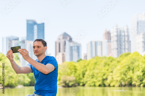 Plakat Młody człowiek siedzi przy selfie w Piedmont Park w Atlancie, w stanie Georgia z malowniczą wodą i panoramę miasta miejskich drapaczy chmur w centrum, Lake Clara Meer