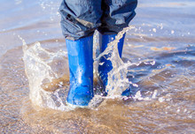 Freeze Action  Of Child Legs In Blue Rubber Boots Jumping Over A Puddle Splashing Water After Rain At Spring,  Summer Or Autumn Day,  Show Water Droplets