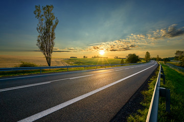 Wall Mural - empty asphalt road around farm fields in rural landscape at sunset