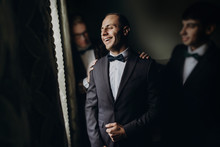 Stylish Groom Laughing And Having Fun With Groomsmen While Getting Ready In The Morning For Wedding Ceremony. Luxury Man In Suit With His Friends Smiling In Room. Space For Text.