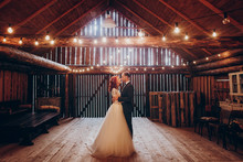 Stylish Groom And Happy Bride Hugging Under Retro Bulbs Lights In Wooden Barn. Rustic Wedding Concept, Space For Text. Newlyweds Couple Embracing, Sensual Romantic Moment
