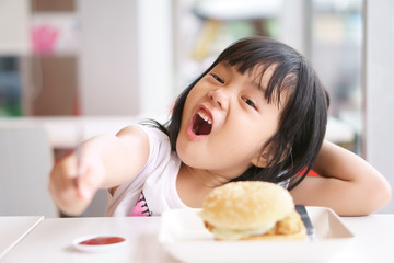 Wall Mural - Asian children cute or kid girl delicious eating ham burger or junk food with tomato sauce or ketchup on white dish and table for breakfast or lunch with hold knife for happy fun smile at restaurant