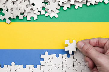 Gabon flag  is depicted on a table on which the human hand folds a puzzle of white color