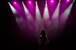 © nagaets - Girl in long gown performing on stage. Girl singing on the stage in front of the lights. Silhouette of singer standing on stage at microphone.