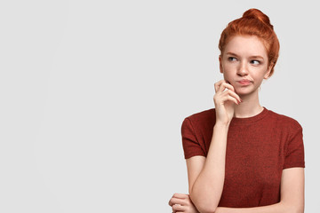 Horizontal portrait of freckled red haired female holds chin, has some intention in mind, feels puzzled and uncertain, looks thoughtfully aside, isolated on white wall with copy space for promotion