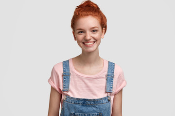 Wall Mural - Happy young female college student laughs happily and looks directly at camera, being in high spirit as has summer holidays, wears denim overalls and t shirt, poses against white background.