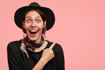 Wall Mural - Horizontal portrait of overjoyed beautiful female wears cowboy style of clothes, indicates at upper right corner with fore finger, stares at camera, isolated over pink background, advertises something