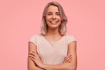 Wall Mural - Horizontal shot of pretty successful young female blogger, keeps hands crossed, glad to achieve success, has dyed hair, wears braces on teeth and casual clothing, isolated on pink background