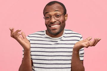 Happy hesitant African American male with cheerful expression, shrugs shoulders in bewilderment, can`t make decision, looks clueless, wears casual striped t shirt, isolated on pink background