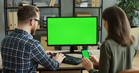Back view on the Caucasian man taping on the laptop computer and woman next to him scrolling on the smartphone at the office table. Green screen. Chroma key. Indoors