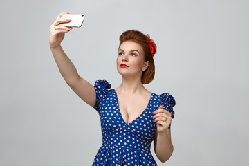 Wall Mural - Waist up portrait of gorgeous fashionable young lady dressed like 1950s pin up girl holding smart phone above her and taking selfie while posting pictures via social networks, having confident look