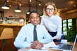 © pressmaster - Two intercultural associates looking at camera while having meeting by table in cafe