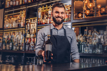 Cheerful stylish brutal bartender in a shirt and apron presents a bottle of exclusive alcohol at bar counter background.