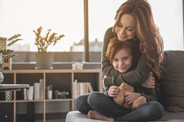 Perfect day together. Portrait of caring mother hugging her daughter at home. They are showing their affection with smile and joy. Copy space in left side