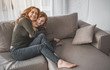 Love you so much. Full length portrait of delighted red-haired woman hugging her daughter tightly on couch. Little girl is holding her teddy bear with smile and joy